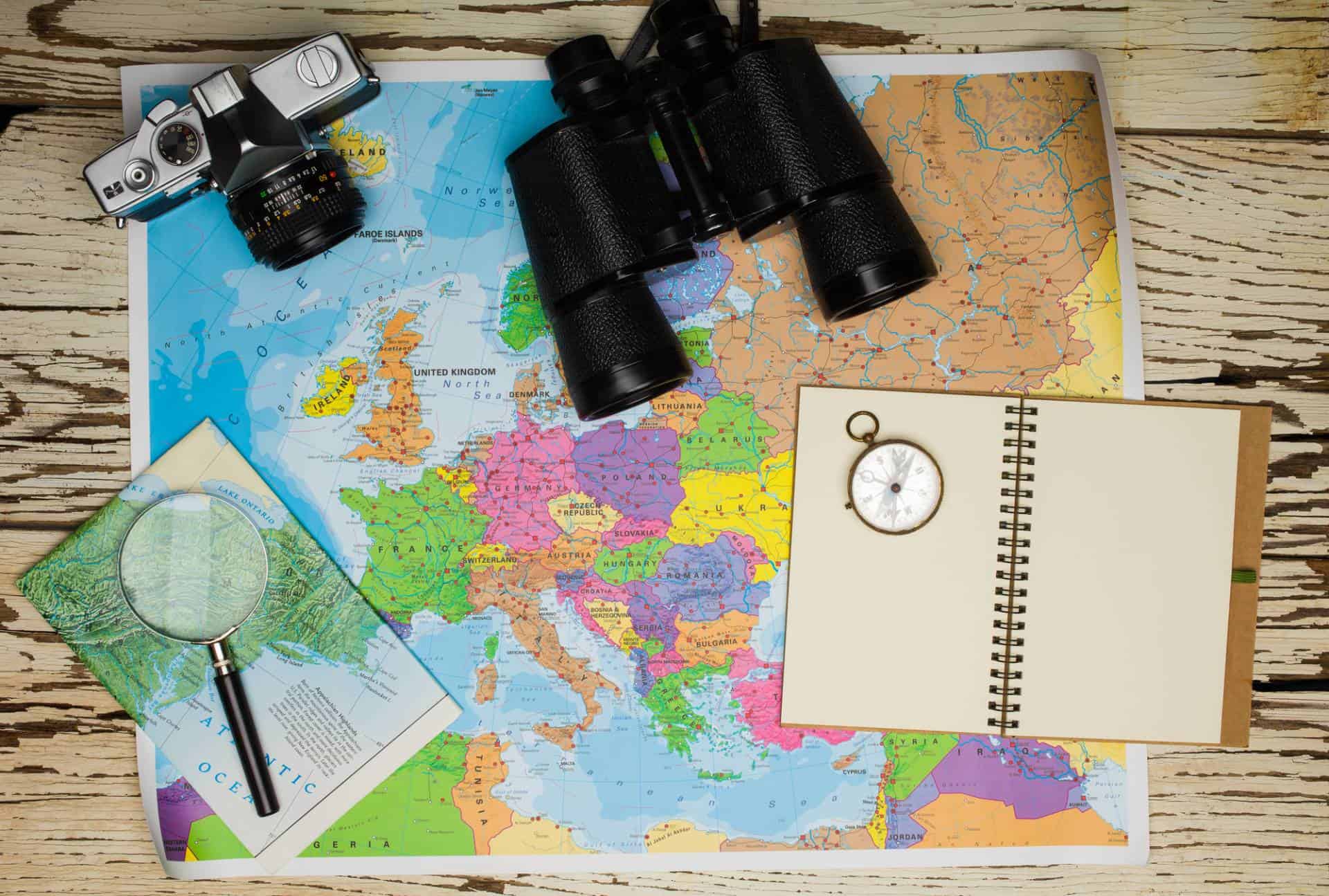 Top view of notebook, binoculars, camera, magnifying glass, pocket watch and map of Europe.