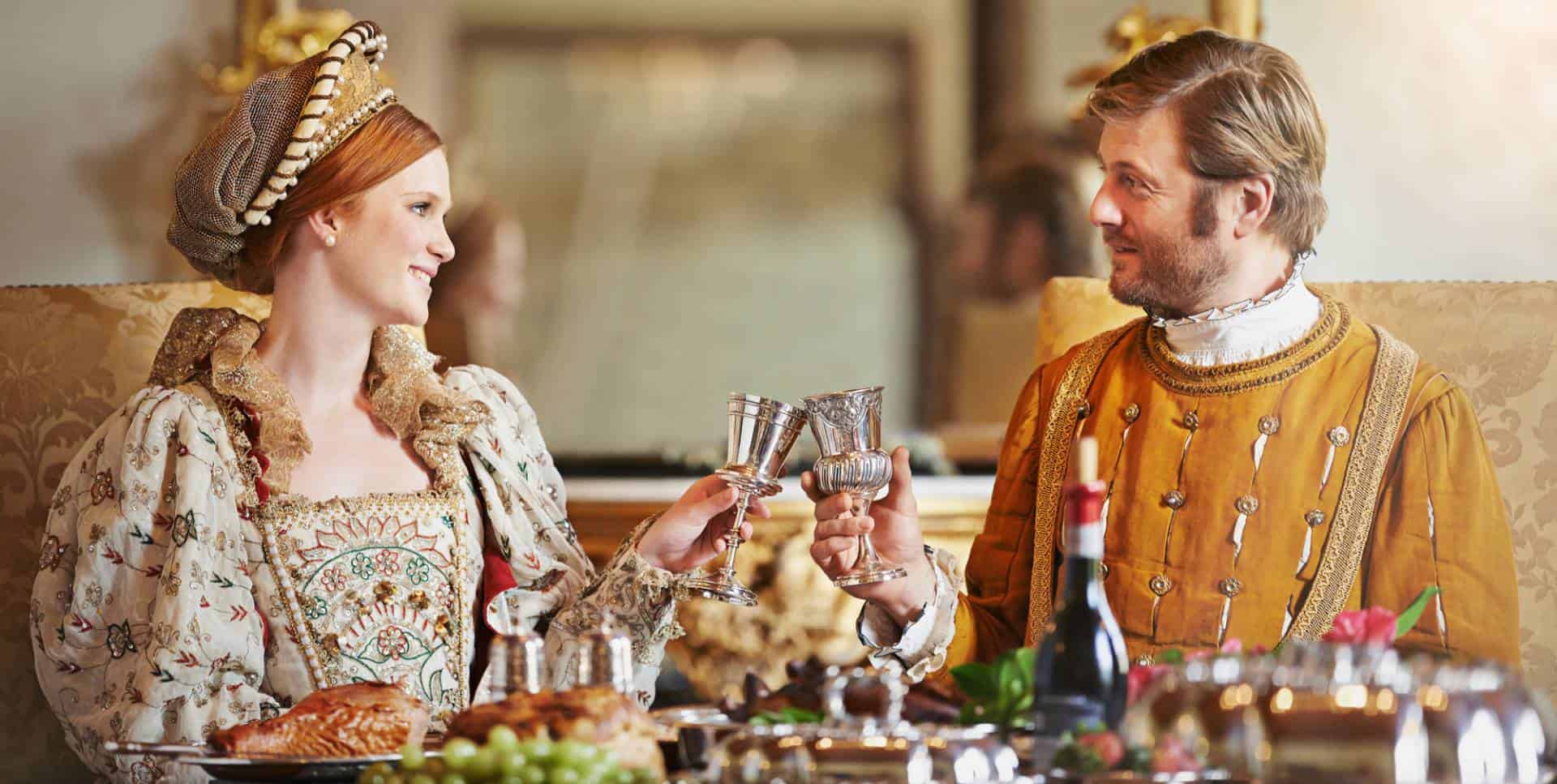 Man and woman wearing costumes of the nobility toasting at a dining table filled with a feast.