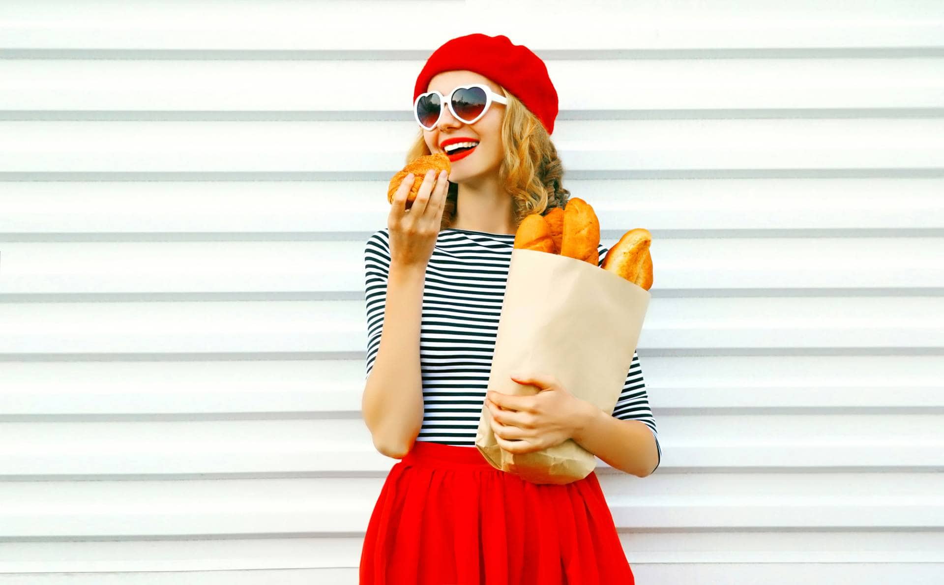 Pretty woman eating croissant, wearing red beret and sunglasses and holding paper bag of baguettes.