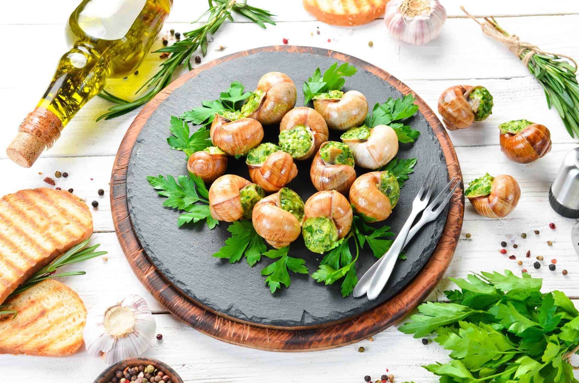 Baked escargot with garlic butter, fresh herbs on a black plate, on white wooden table.