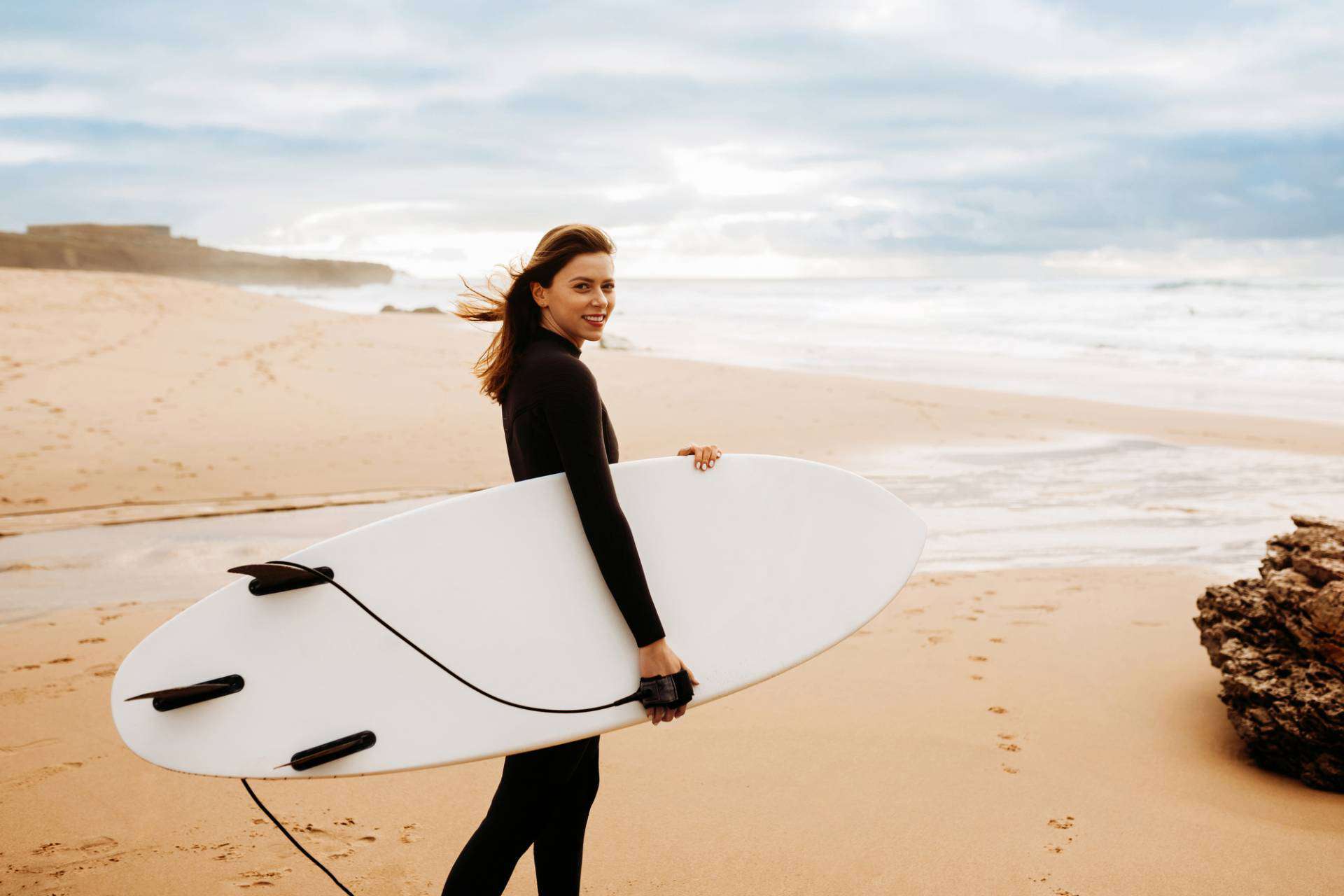 Young woman in wetsuit walking on beach with surfboard looking and smiling at camera.