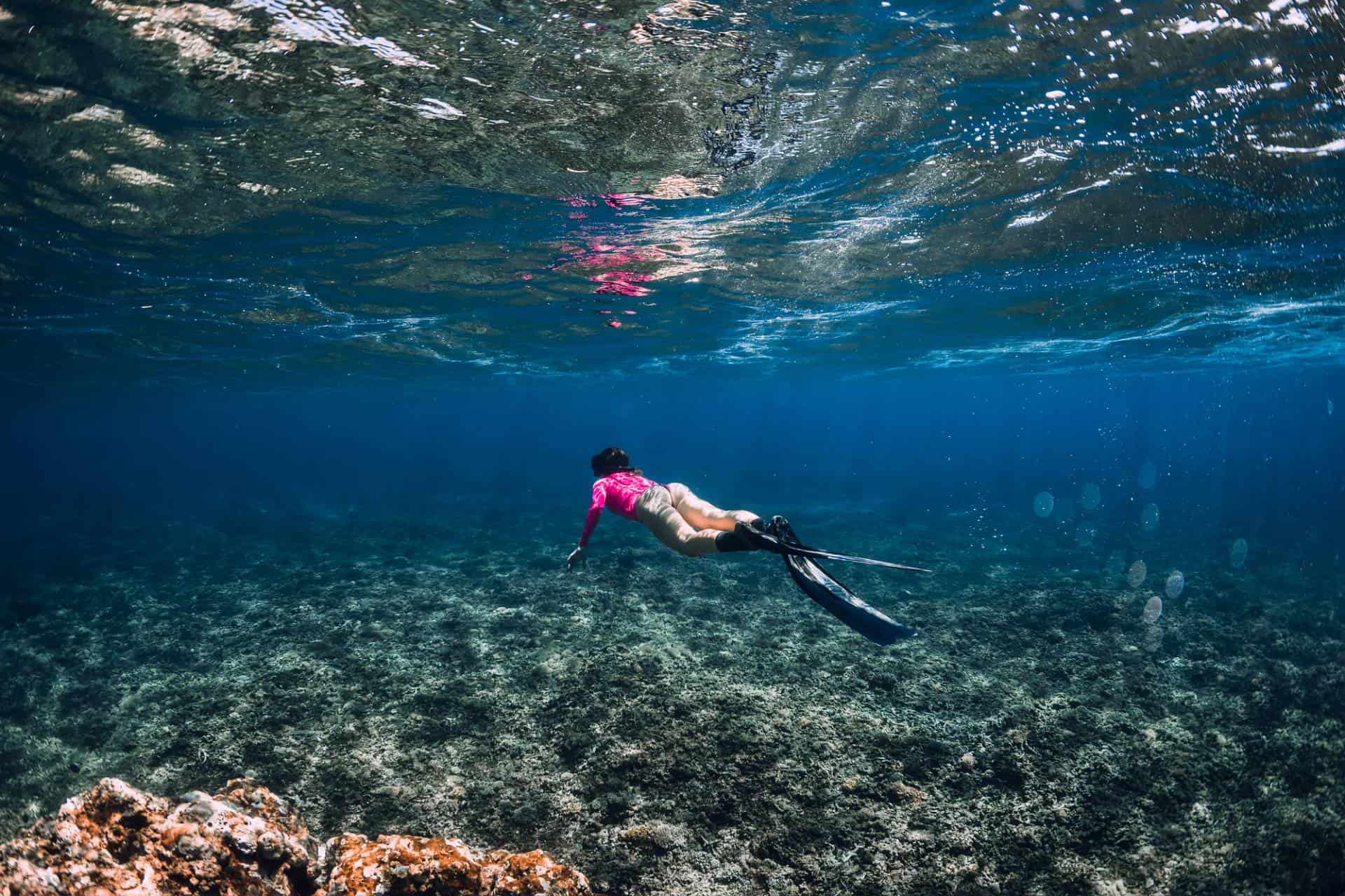 Female diver in pink suit glides over coral reef in clear blue sea.