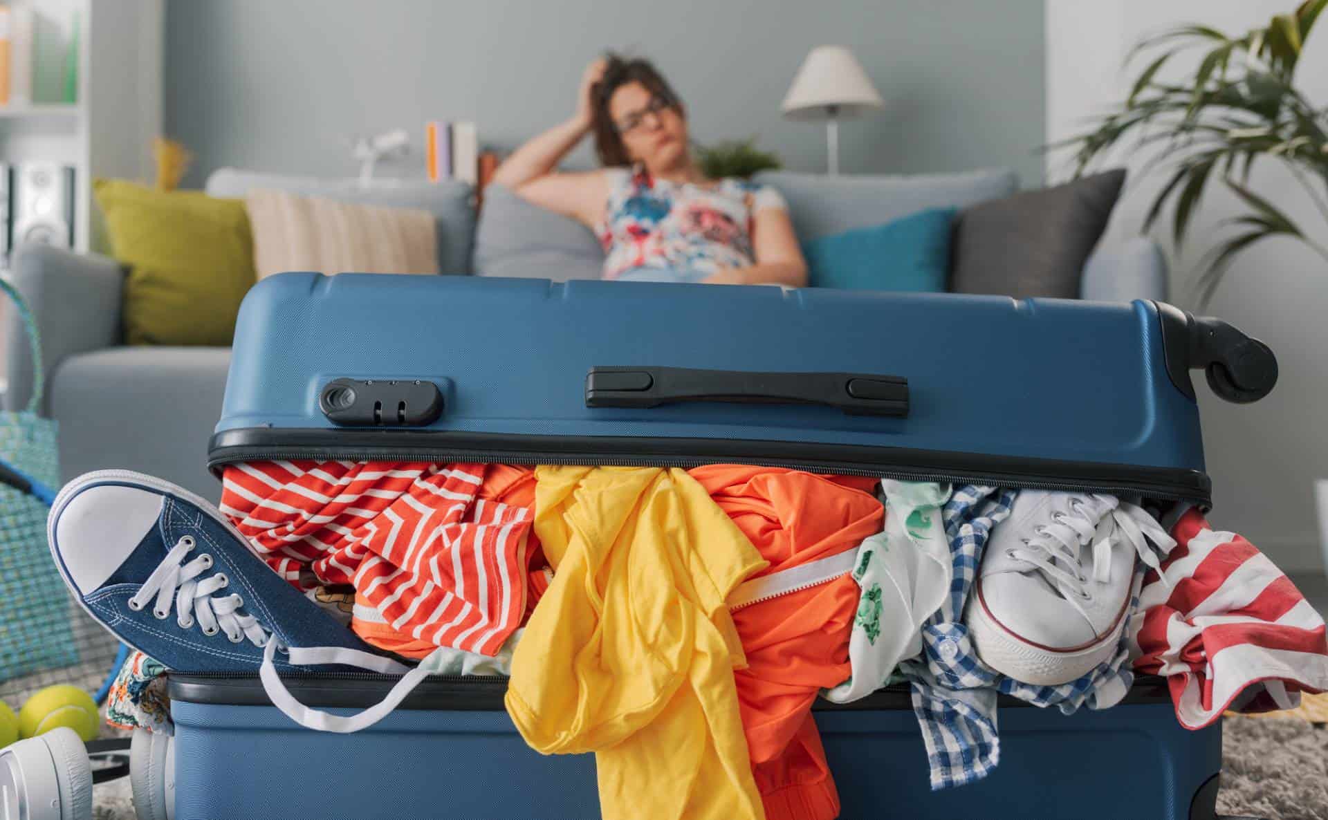 Stressed woman sitting on couch behind overpacked suitcase.